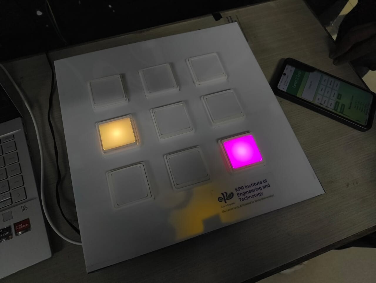The NeuroGlow interactive rehabilitation system, showing a grid of square response panels, two illuminated in yellow and magenta, beside a smartphone displaying the system interface.